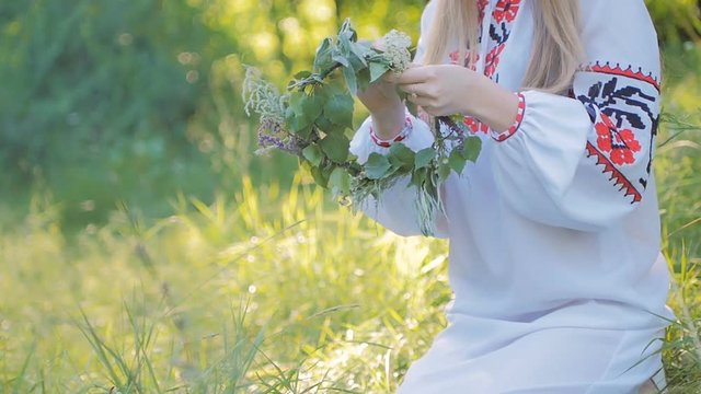 Slavic woman in an embroidered shirt patterns , weaves a wreath of wild flowers sitting by the fire .