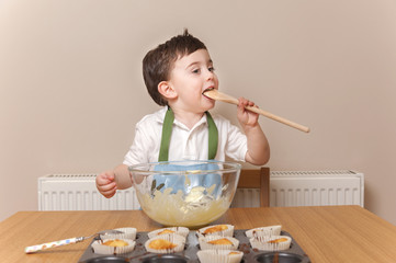 Cute young boy inside the kitchen and licking a wooden spoon. He is baking cupcakes and he has been...