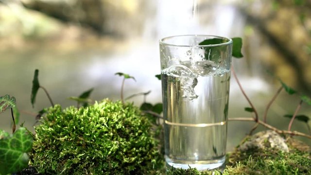Glass of drinking natural water with a waterfall in soft focus on background. Pure ecological water from a spring in the mountains. Water pours in the glass standing on mossy rock.
