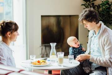 Young happy family with newborn baby boy having breakfast in cafeteria. Dad holding little son while mom eating her breakfast. People, healthy lifestyle, family and food concept