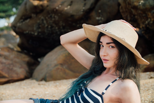 Long haired girl In a hipster hat and striped swimsuit relax on tropical beach, background of stones.