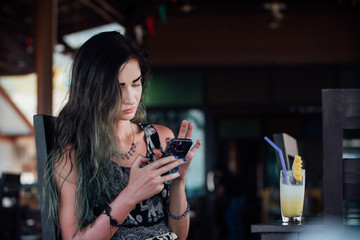 The girl typing in the phone, sitting in a cafe with fruit pineapple juice in a glass and a tube.