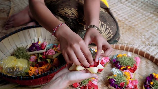 Close up of balinese women making offerings (Canang sari) - putting different flowers on canang (small palm-leaf basket as tray) sitting on bamboo mat. Shot with Sony a7s on slider.