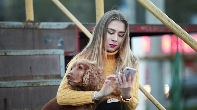 Blonde girl and dog cocker spaniel Adjusts her hair looking at the reflection on smartphone looking for information on the Internet in the evening city Against the background of metallic rusty steps.