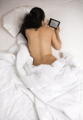 Woman with naked back lying in bed and reading an ebook