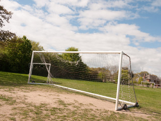 An Empty and Unused Goal Post with A White Net in the Middle of A Park with Grass and Soil on the Ground, Wheels on the Frame to Help Move it and Houses and Trees in the Background