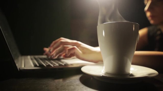 Still shot of a woman's hands typing on a laptop keyboard, with a cup of hot coffee near.