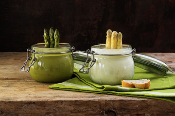 two asparagus cream soups white and green in glass jars on a rustic wooden board, dark background,...