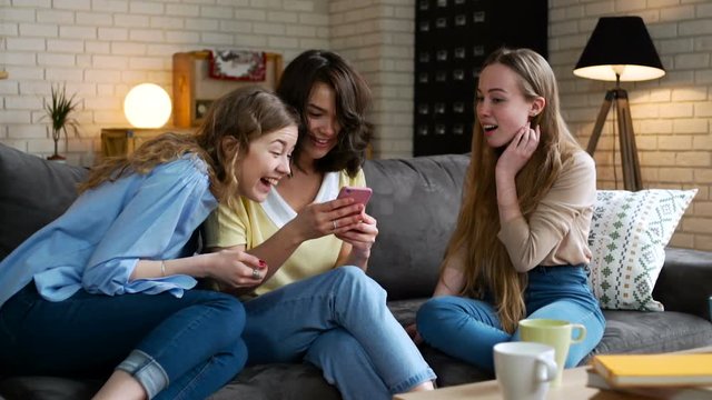 three young and energetic best friend girls look over photos in social network using telephone mobile and discuss emotionally, laughing and impressing surprise sitting on sofa at home during sunny day