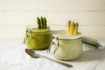 white and green asparagus cream soup in glass jars against a bright wooden background with copy...