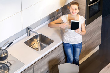 Beautiful young woman holding tablet in the kitchen and showing screen with finger and toothy smile looking at camera.