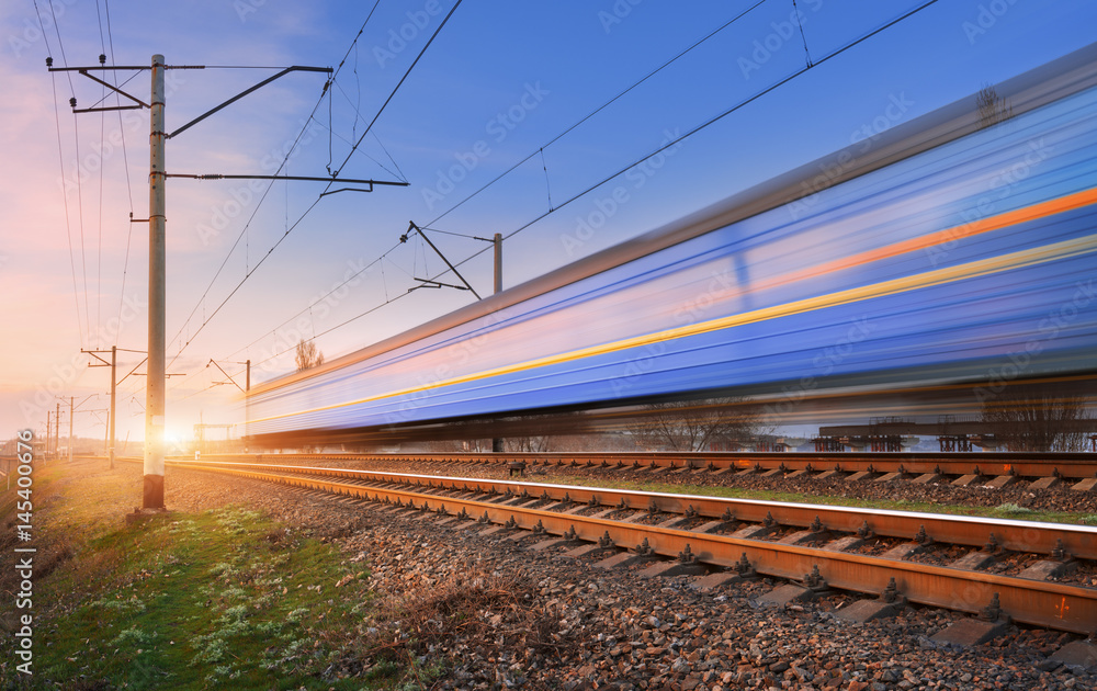 Wall mural High speed passenger train in motion on railroad at sunset. Blurred commuter train. Railway station against colorful blue sky. Railroad travel, railway tourism. Rural industrial landscape. Concept