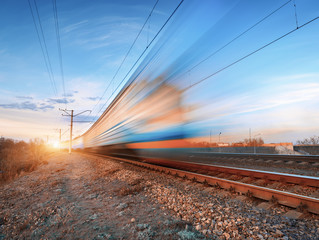 High speed train in motion on railroad track at sunset. Blurred commuter train. Railway station against colorful blue sky. Railroad travel, railway tourism. Rural industrial landscape in twilight