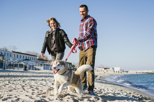 Young caucasian couple walking on sea beach with siberian husky dog