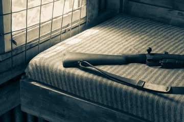 Light through side window security bars on bed with striped cover and rifle on hunters shed bed