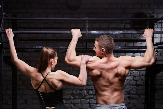 Rear view photo of muscular man and woman doing exercises on horizontal bar against brick wall at the cross fit gym.
