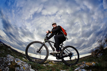 Cyclist in the black sportwear riding the bike on the rock at evening against beautiful blue sky with clouds.
