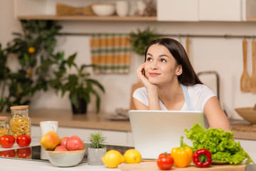 Obraz na płótnie Canvas Young woman standing near desk and laptop in the kitchen, smiling, dreaming. Food blogger concept