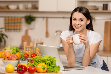 Young woman standing near desk and laptop in the kitchen, smiling, with cup of tea