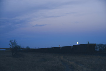Train Sits Near Rural Highway on Plains at Dusk with Moon Rising in Colorful Pastel Sky 