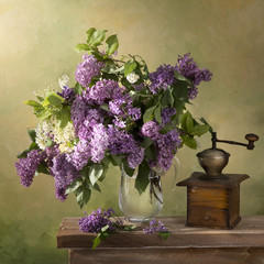 Still life A bouquet of lilac in a glass jug on a wooden table with a picturesque background and an old coffee grinder