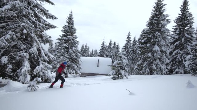 Camp in the winter mountains. Traveler in snowshoes walks next to a cabin covered with snow, in a coniferous forest. Winter adventures in a mountains.