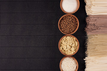 Decorative border of bundles raw noodles with ingredient in wooden bowls on black striped mat background with copy space, top view.