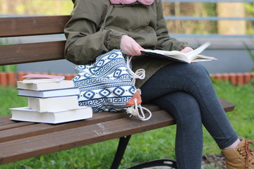Młoda dziewczyna, uczennica, w zielonej kurtce, jeansowych spodniach, siedzi na ławce w parku, na kolanach trzyma rozłożona książkę, obok niej na ławce leży młodzieżowa torba i stos książek  - obrazy, fototapety, plakaty
