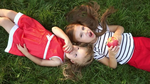 Two little sister girls eating apples lying on green grass in a park. Have a bite in the open air. Family picnic. Happy childhood.