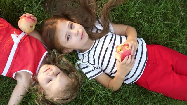 Two little girls sister eating apples lying on green grass in the park. Snack outdoors. Family picnic. Happy childhood.