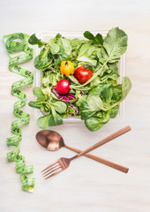 Fitness dieting salad with cutlery and measuring tape on white wooden background, top view. Mixed greens salad bowl for healthy  lunch.