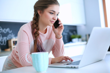 Young woman sits at the kitchen table using a laptop and talking on a cell phone