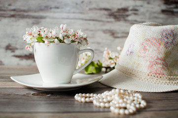Obraz na płótnie Canvas Cup of blooming flowers pear tree, beads from pearls, wicker hat on a gray wooden background.