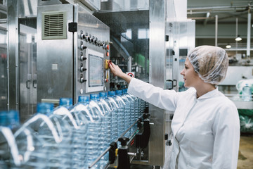 Young happy woman worker checking robotic line for bottling and packaging pure drinking water into...
