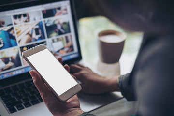 Mockup image of a business woman holding mobile phone with blank white screen while using laptop with coffee cup on the table in office
