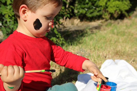 niño pequeño pintando con los dedos en el jardín con la cara manchada de pintura