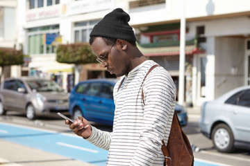 Fashionable young black man tourist with leather backpack looking at smartphone in his hands with serious expression, using online navigation app, searching for direction while got lost in big city