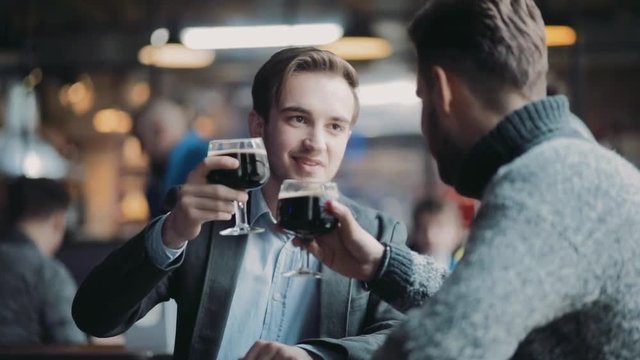 Two young guys having fun in the pub, toasting and drinking lager beer by the bar. Pub interior, visitors on the background. Leisure time, being happy. Friends forever, alcohol mood.