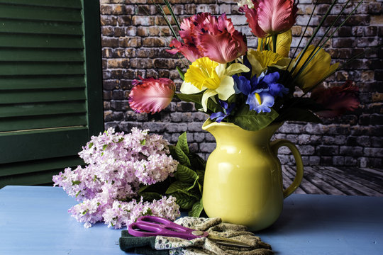 fresh cut spring flower bouquet in yellow water pitcher on blue table with clippers and work gloves with green shutter and brick wall background