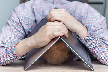 Upset young man covering his head with his laptop. Burn out at work concept.