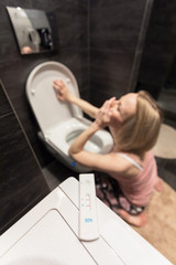 Pregnancy test in the foreground and sad sick woman sitting on her knees over the toilet in the bathroom. Toxicosis. The first signs of pregnancy