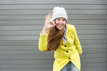 Smiling brunette girl in yellow coat shows ok sign against gray background.