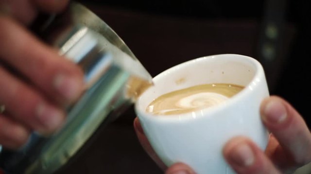 Barista pours milk into a cup of espresso, mixing forms cappuccino, paints on milk foam. Close-up.