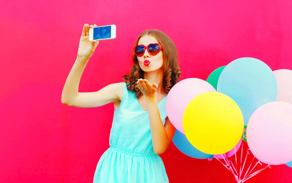 Fashion pretty woman taking a picture on a smartphone sends an air kiss over an air colorful balloons a pink background