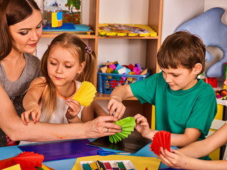 Preschool scissors in kids hands cutting paper with teacher in class room. Development and social lerning happy children in school. Couple girl and boy with woman together.