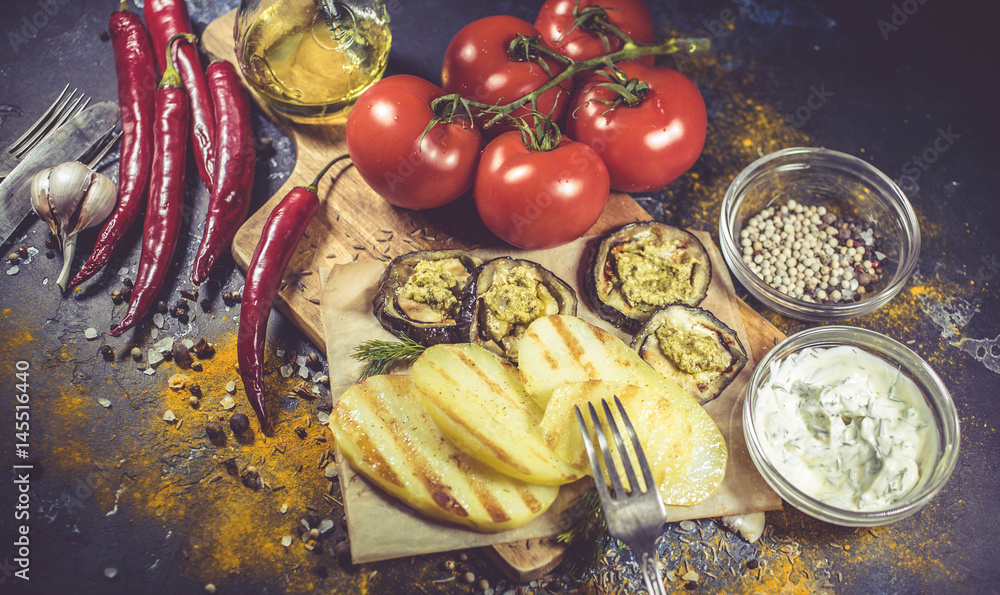 Wall mural Fried slices potatoes and eggplant, fresh red tomatoes, chili pepper, spices on a chopping board on dark background. Selective focus. Food background.