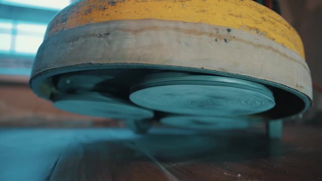 Abrasive discs of yellow floor polishing machine stands down on dusty laminate floor, starts working, slow motion close up