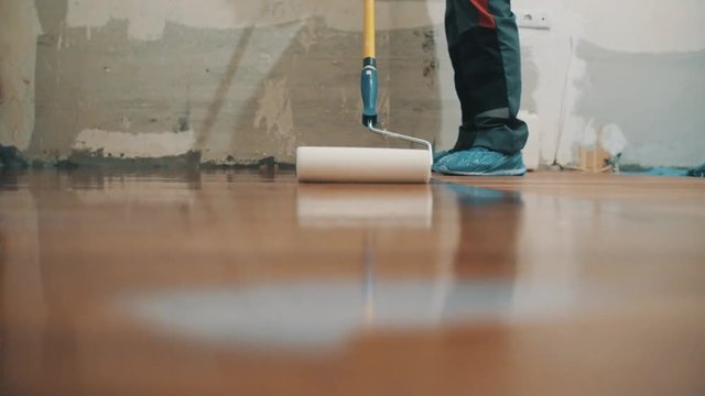 Unrecognizible man in working pants blue shoes applying polisher on new parquet floor using paint roller