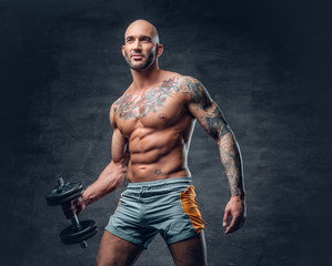 Studio portrait of athletic shirtless shaved head tattooed male dressed in a sports shorts holds...