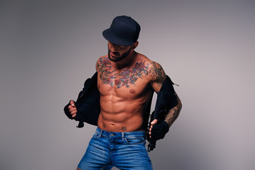 Tattooed male dressed in a jeans, cap and waistcoat on a naked torso.